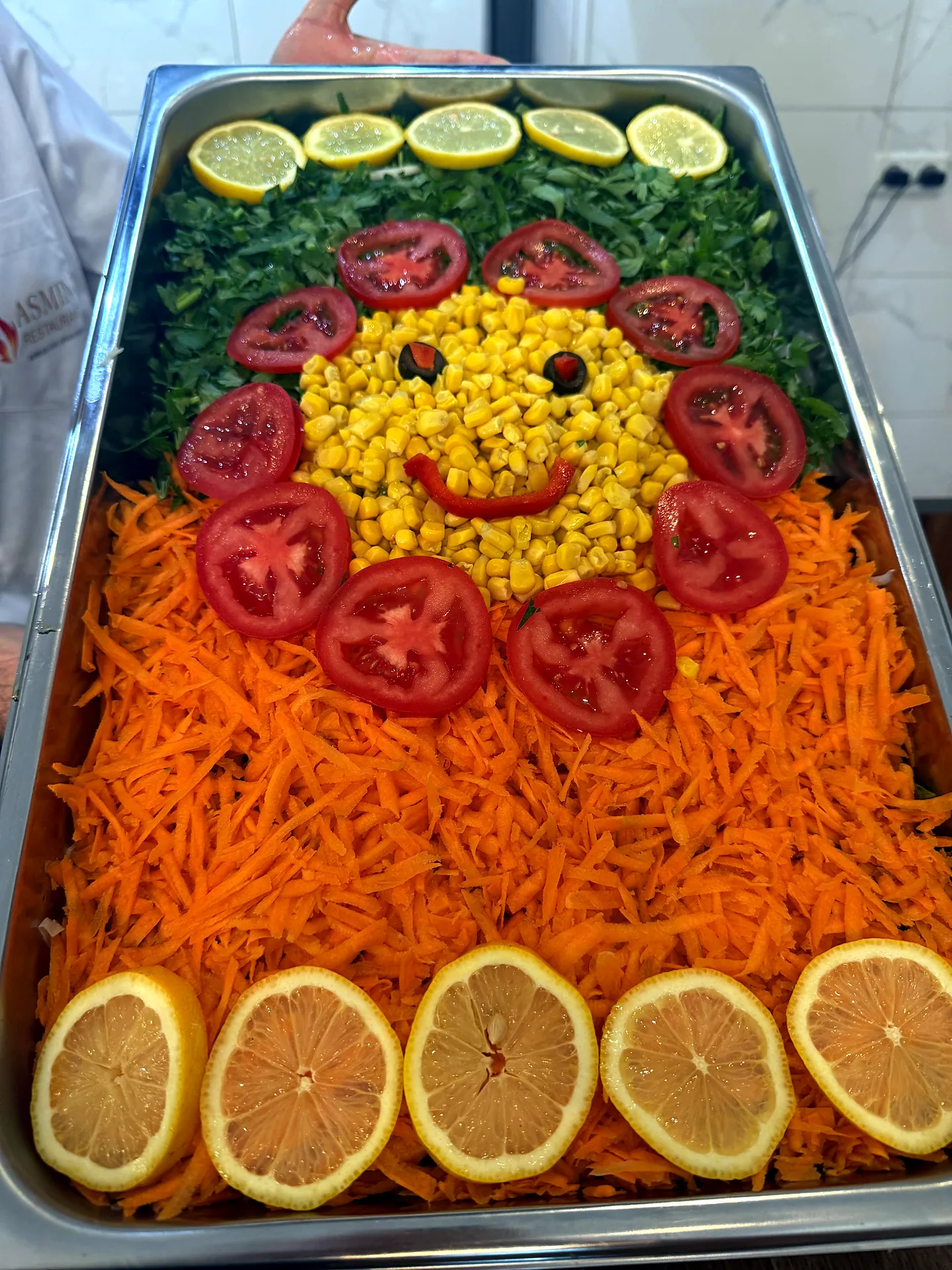 Salatplatte mit Smiley aus Mais und Tomaten, umrahmt von Zitronen und Karotten